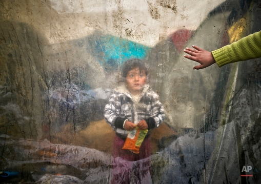 A migrant child stands behind plastic sheets covering tents during a rainfall as a boy waves in a makeshift camp at the northern Greek border post of Idomeni, Greece, Tuesday, March 15, 2016. Hundreds of migrants and refugees walked out Monday of an overcrowded camp on the Greek-Macedonian border, determined to use a dangerous crossing to head north but were returned to Greece. (AP Photo/Vadim Ghirda)